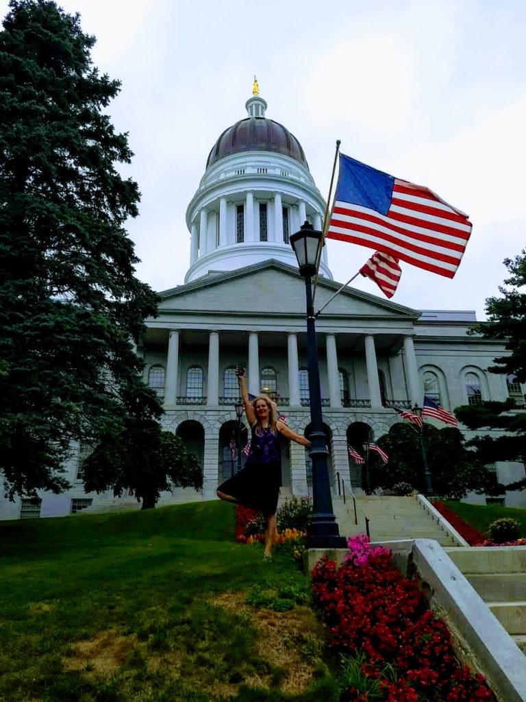 Maine state house with flag and Karen in front. Enjoy the stunning architecture of this state house designed by Bullfinch.