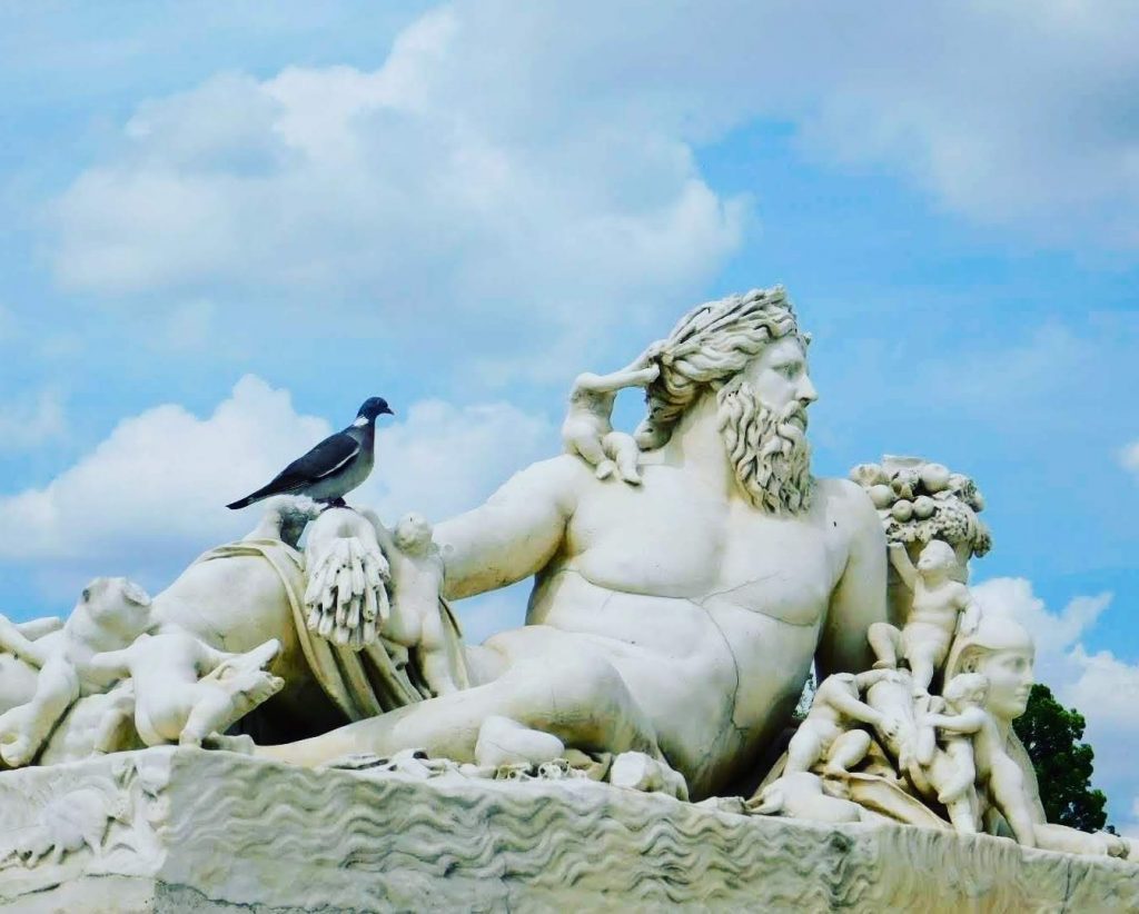 A guy in Paris (statue) relaxing after he beat the shell game. Looks like some god, with cherubs braiding his hair and a rock pidgeon on his arm. Cloudy sky is visible behind him.