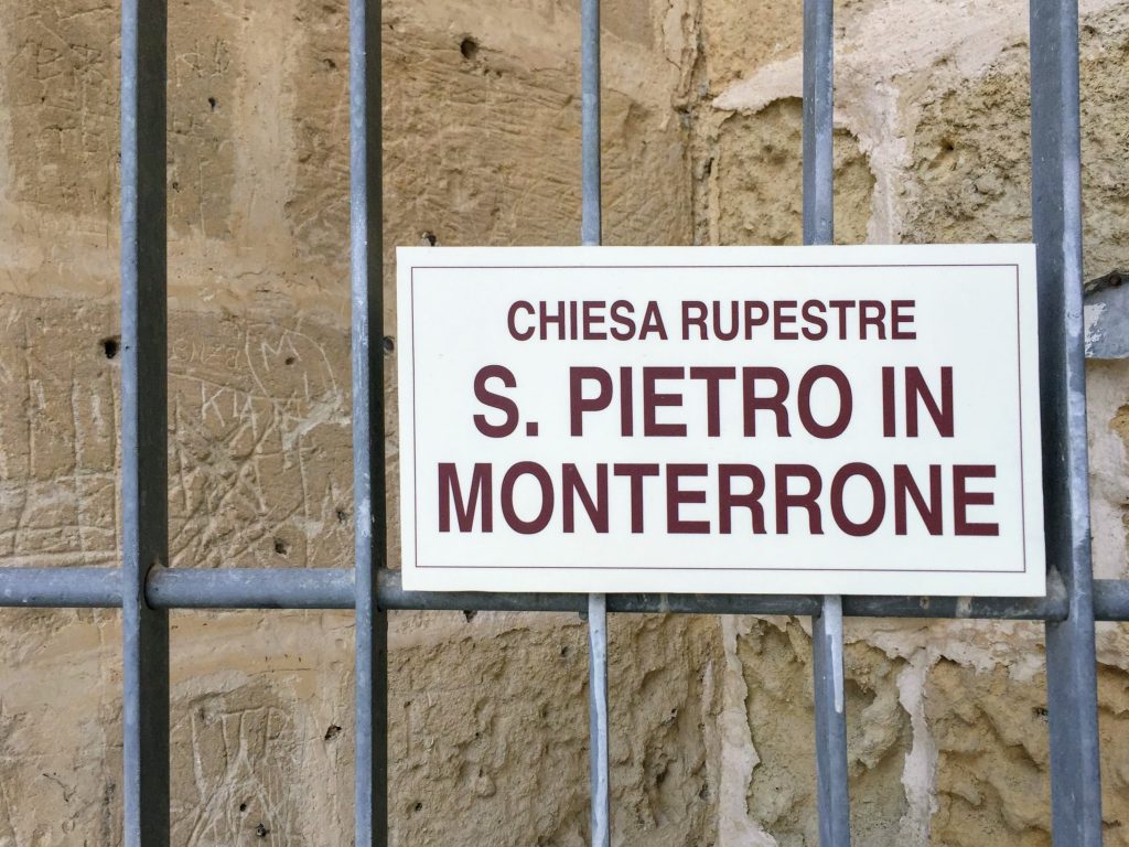 Sign on fence in front of rock reading "Chiesa Repestre: S. Pietro in Monterrone"