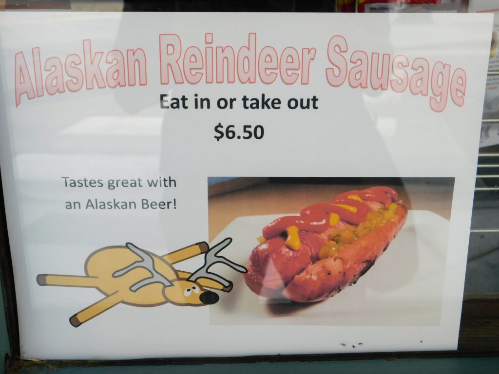 Sign in store window reading "Alaskan Reindeer Sausage" 
"Eat in or take out $6.50"
"Tastes great with an Alaskan Beer!"